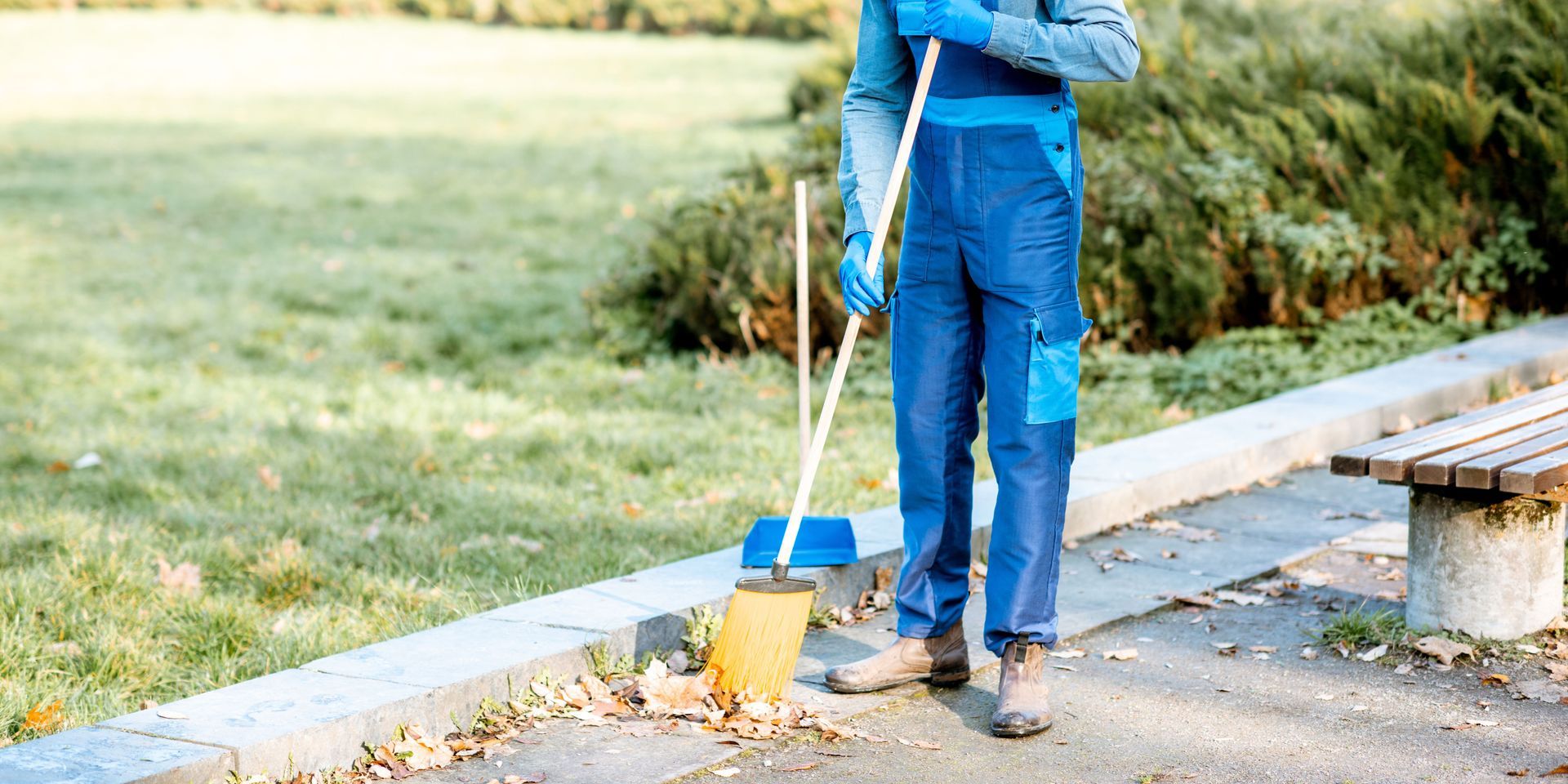 Person in blue overalls sweeping leaves on a sidewalk in a park.