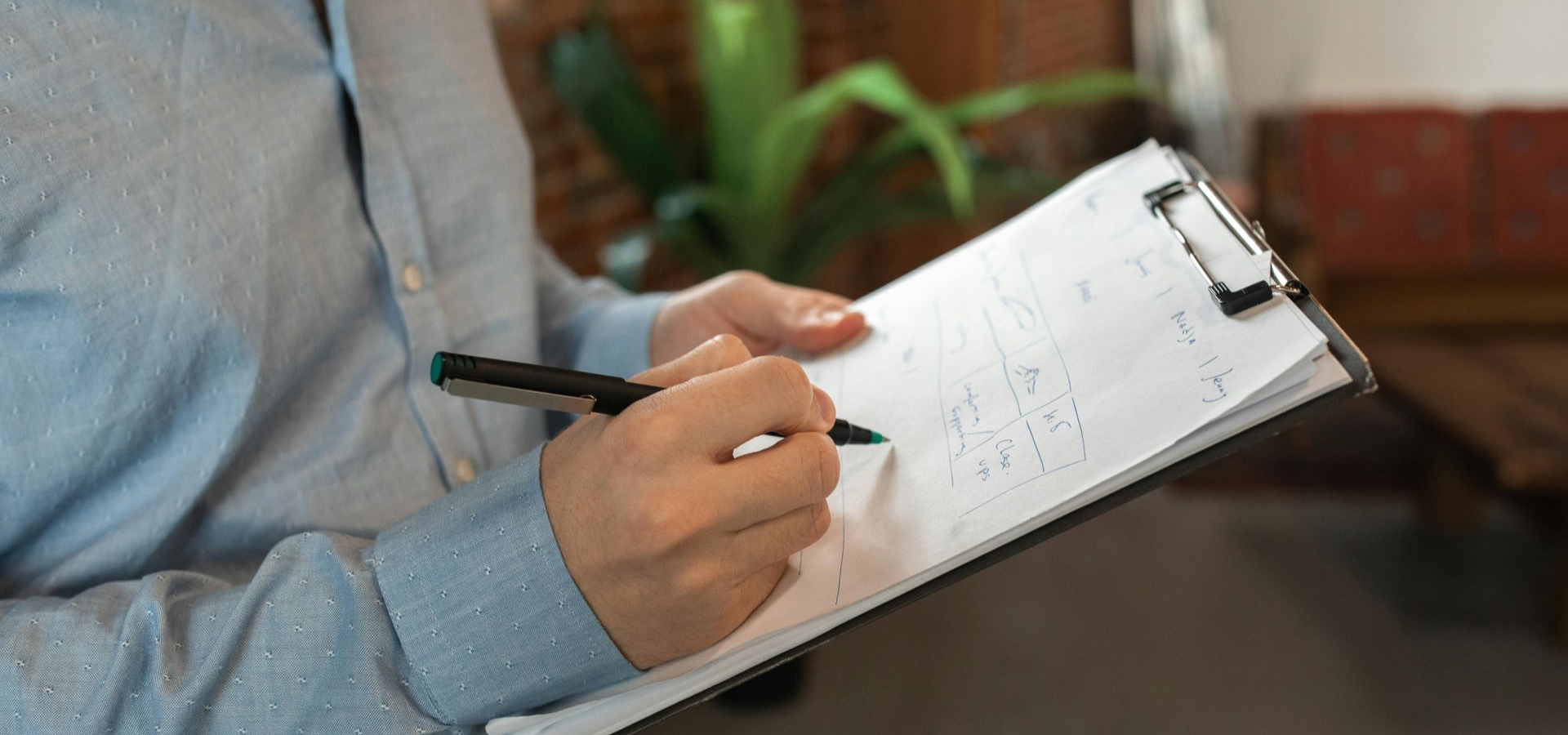 Person in blue shirt writing on a clipboard with a pen, indoor setting with plants and brick wall in the background.