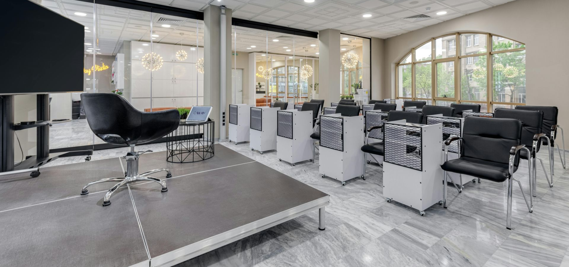 Conference room with rows of chairs, white speakers, and a stage. A large window and a screen on the wall.