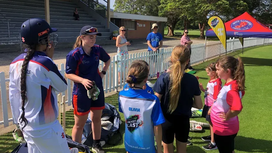 Cricket players in uniform huddle on the field, near equipment and a spectator area.
