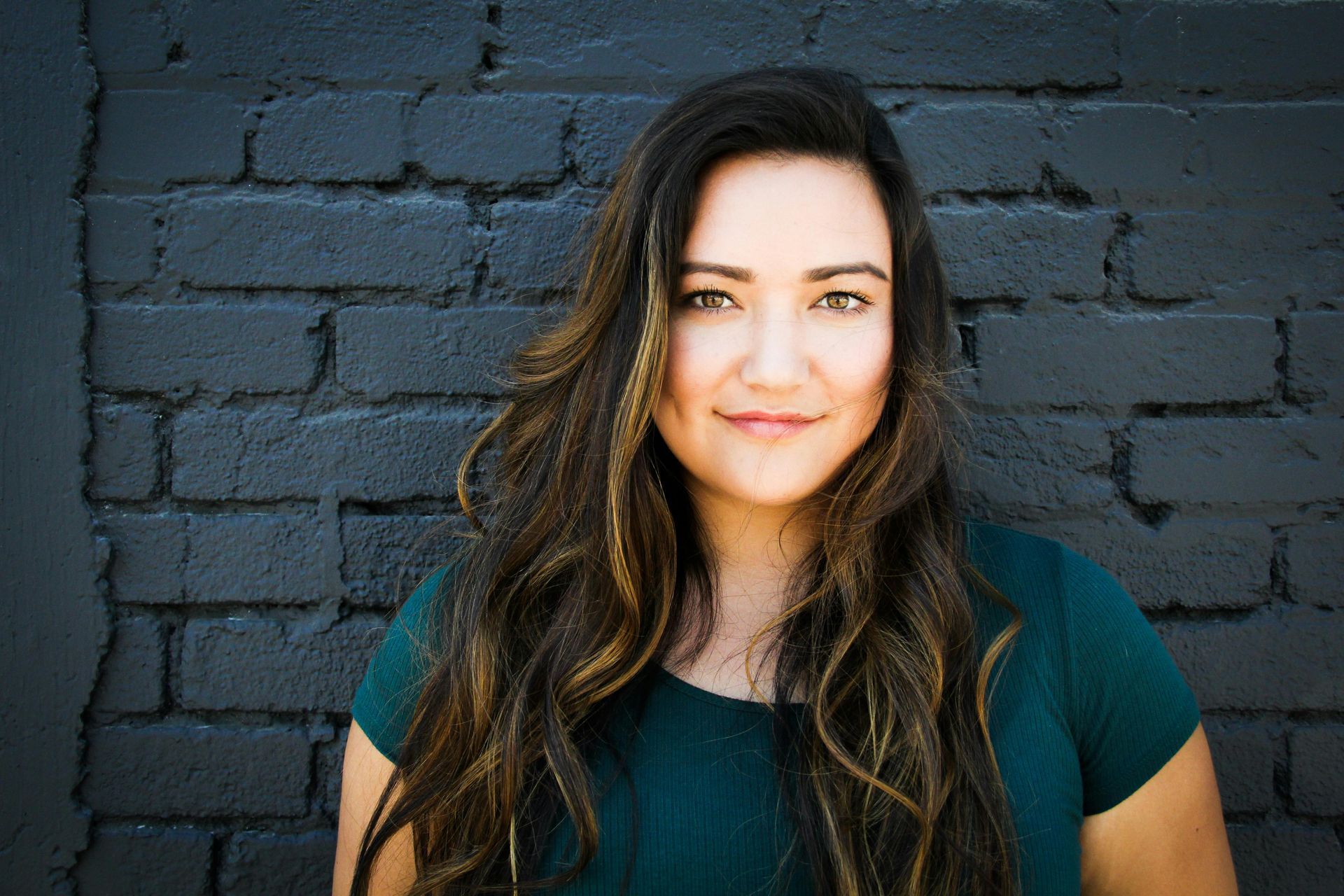 Woman with long, wavy brown hair smiles, leaning against a dark brick wall, wearing a teal top.