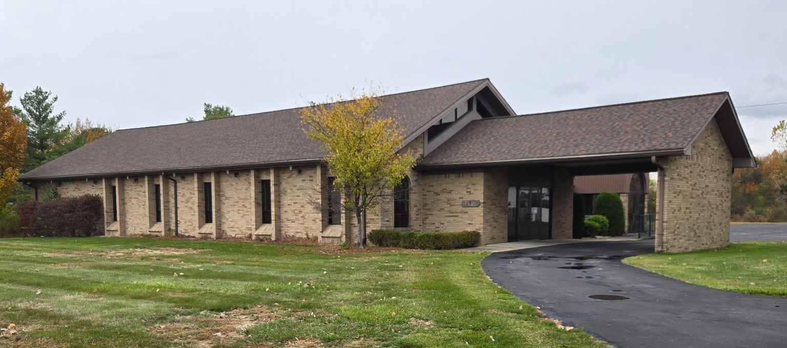 A tan brick building with a brown roof and a black asphalt driveway, surrounded by green grass.
