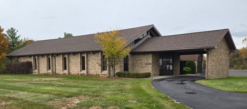 A tan brick building with a brown roof and a black asphalt driveway, surrounded by green grass.