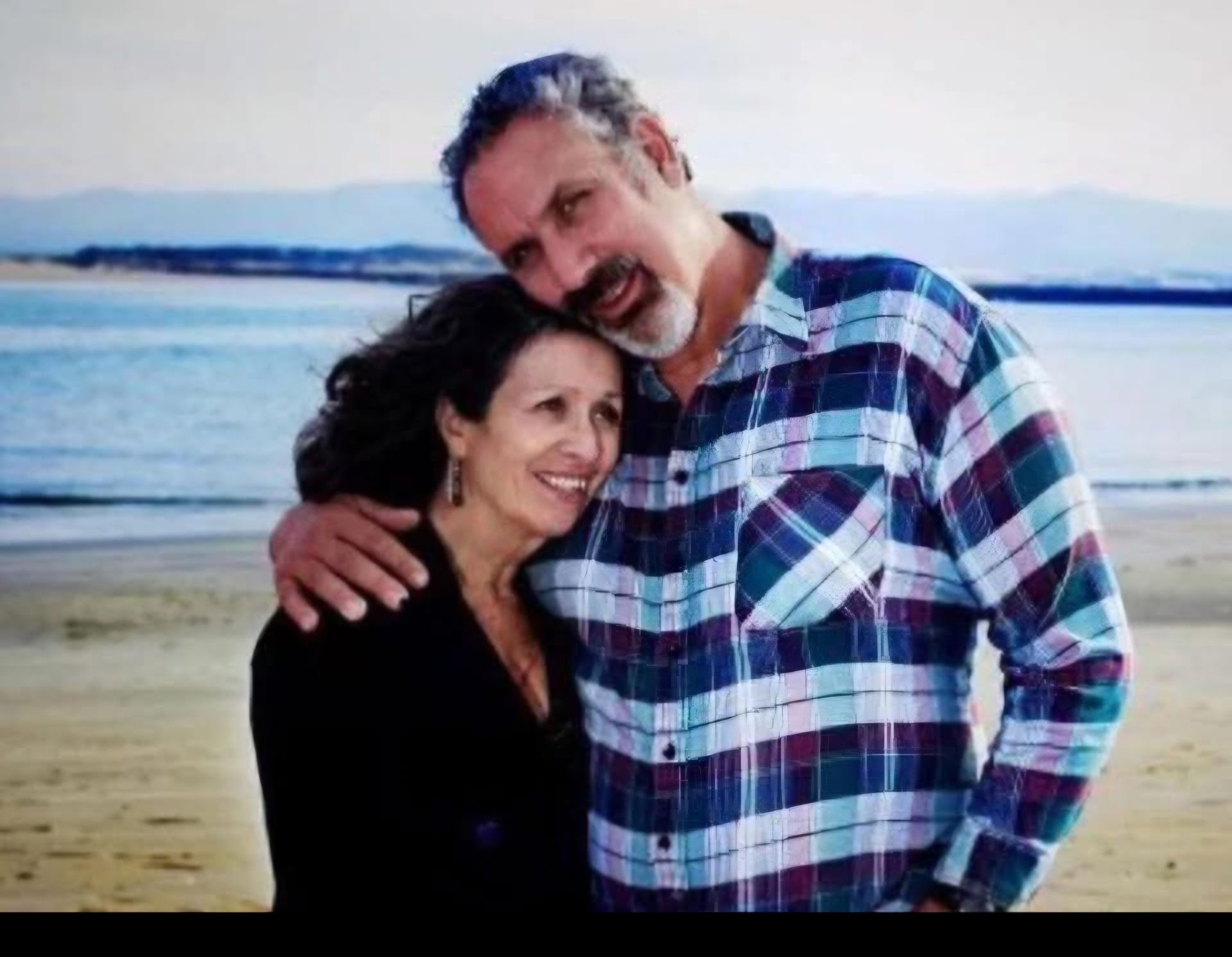 A smiling couple embraces on a beach with a calm sea and distant mountains in the background.