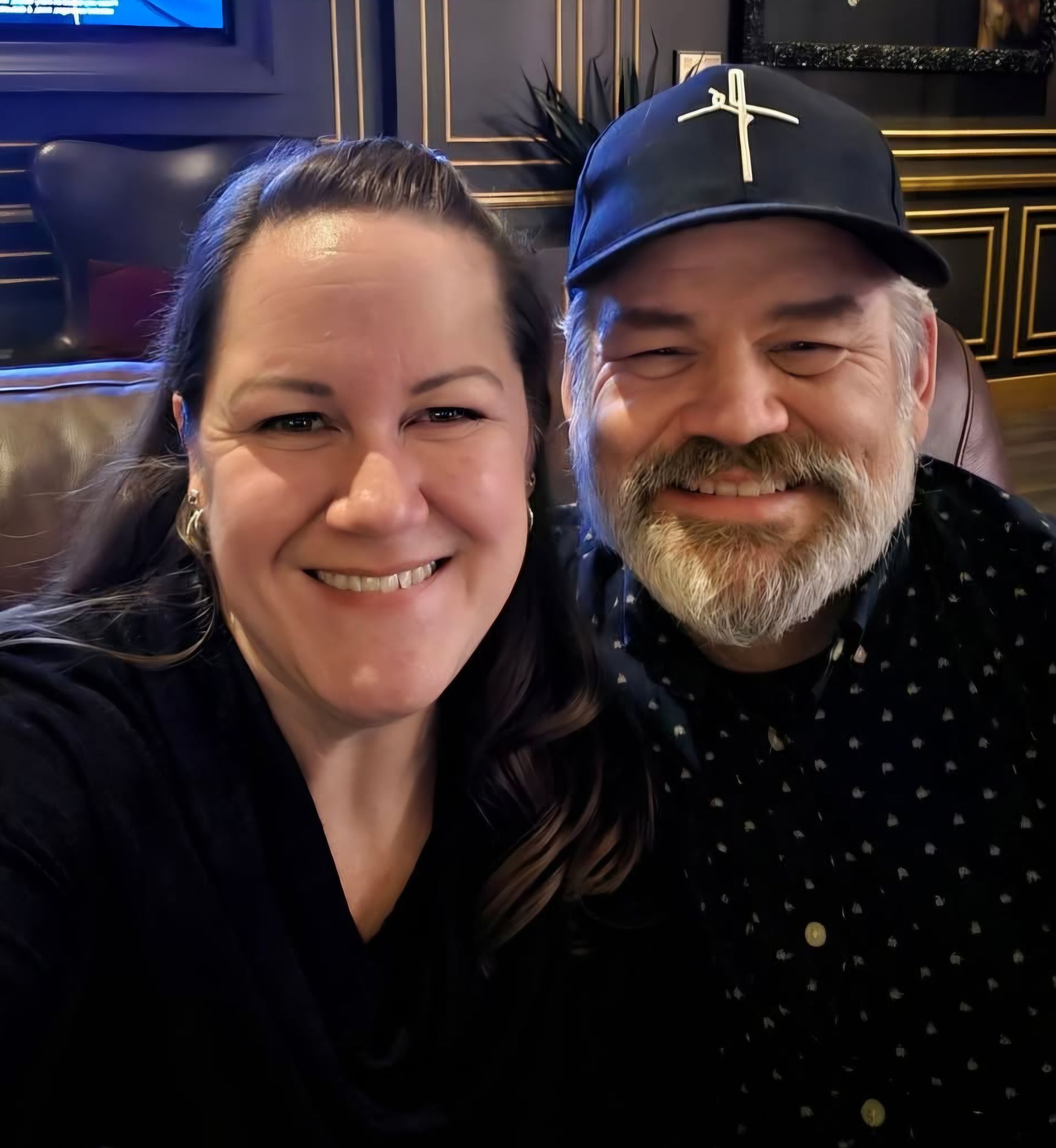 A smiling couple poses for a selfie in an indoor setting with dark walls and gold trim.