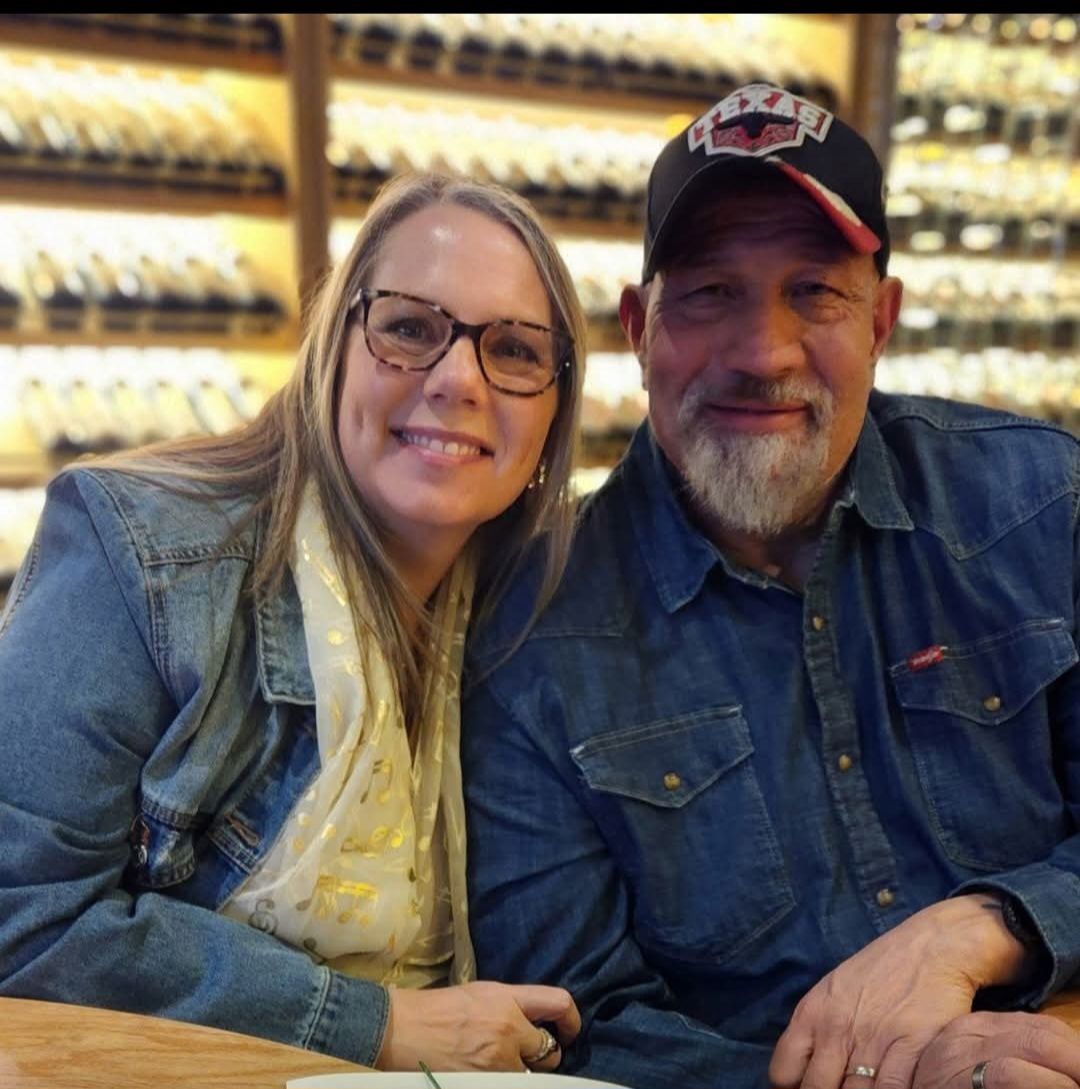 A smiling couple sits together in front of a wine rack, both wearing denim shirts.