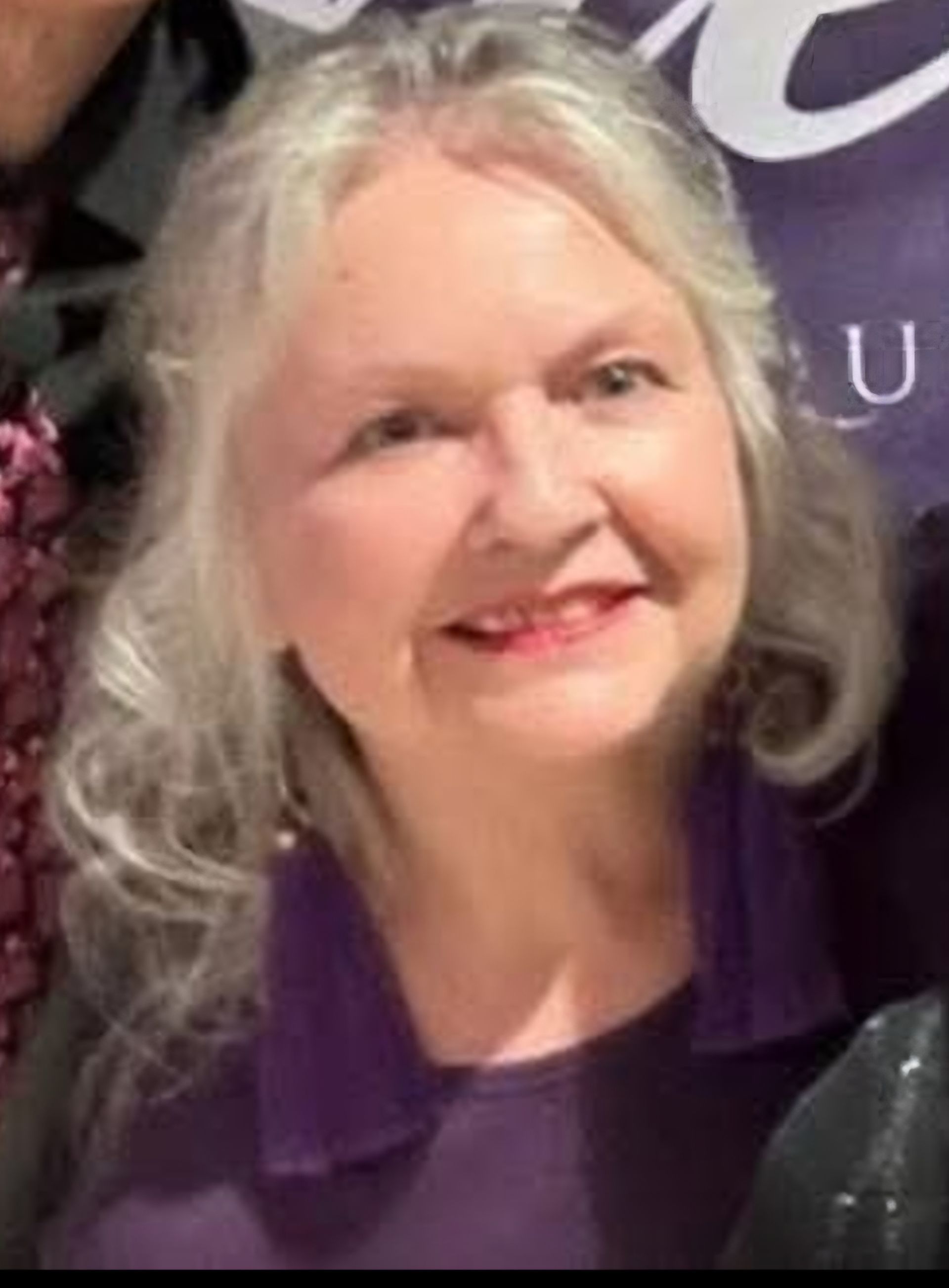 A smiling individual with silver hair, wearing a purple top and long purple earrings, posed against a dark backdrop.