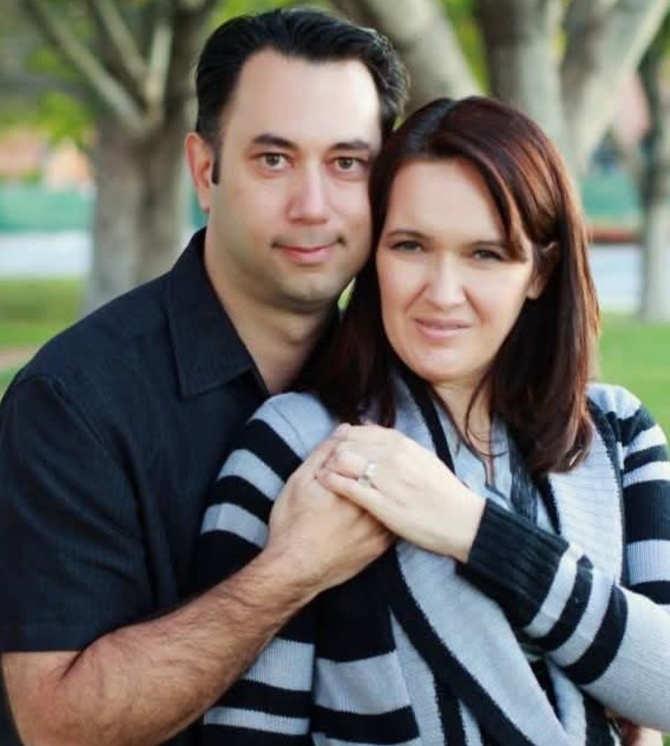 A man stands behind a woman with his arms around her, both smiling in an outdoor setting with trees in the background.