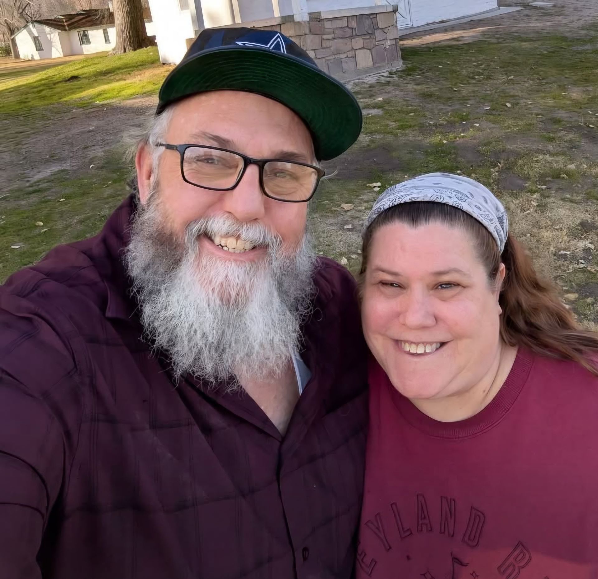 A smiling person with a long beard and glasses wears a baseball cap beside a smiling person in a headband outdoors.