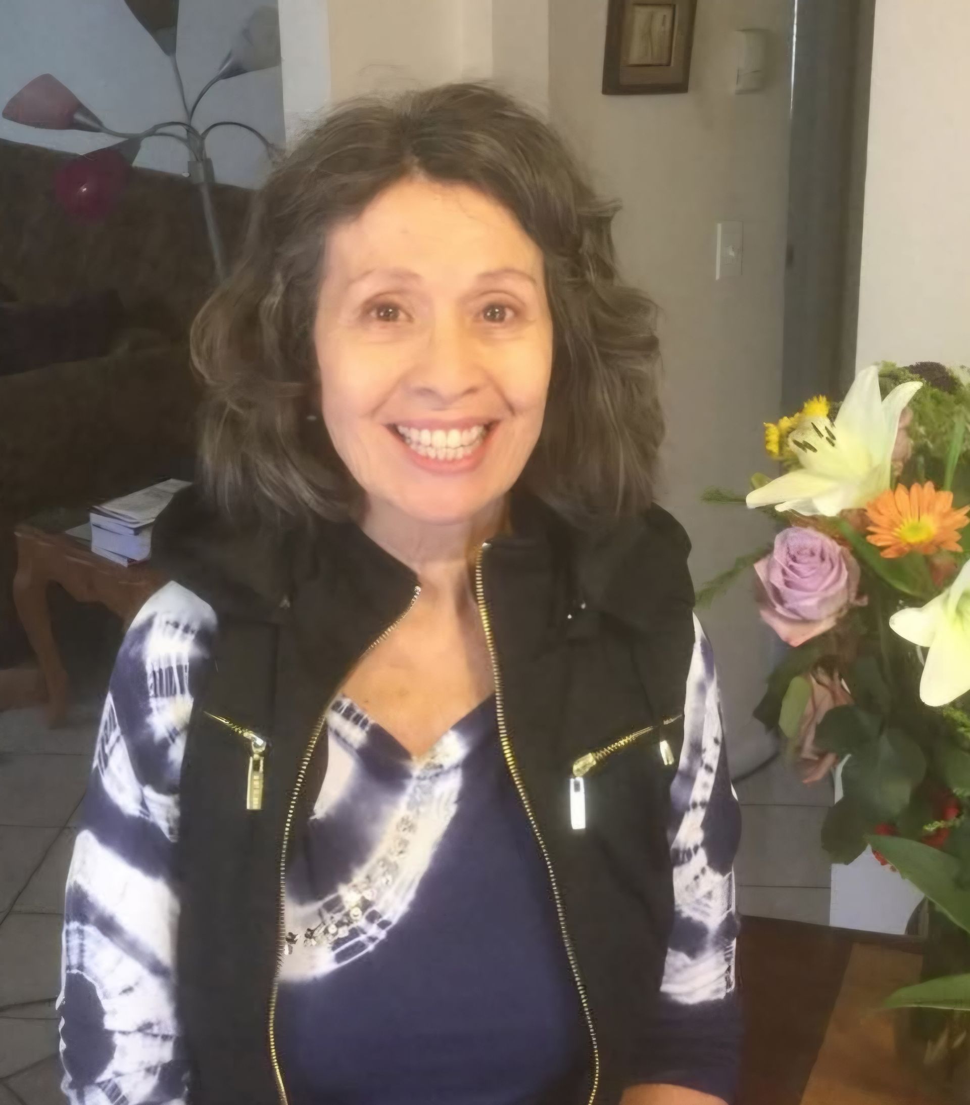 A smiling person in a blue tie-dye shirt and black vest, posing indoors next to a colorful bouquet of flowers.