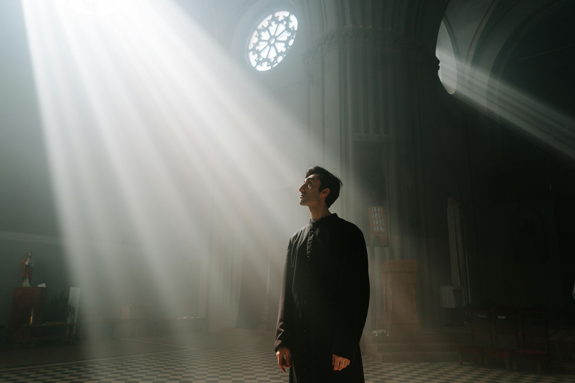 A person stands in a dimly lit cathedral, looking up at a dramatic beam of light streaming through a rose window.