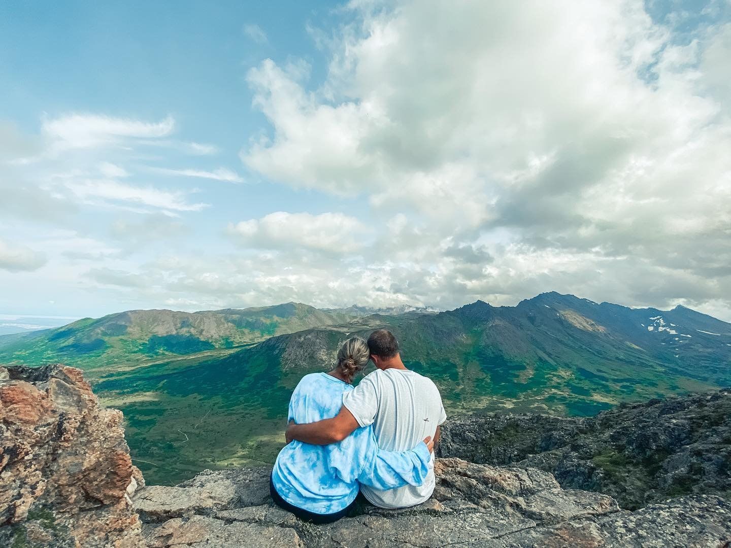 Couple embracing on a mountaintop, overlooking a green valley under a cloudy sky.