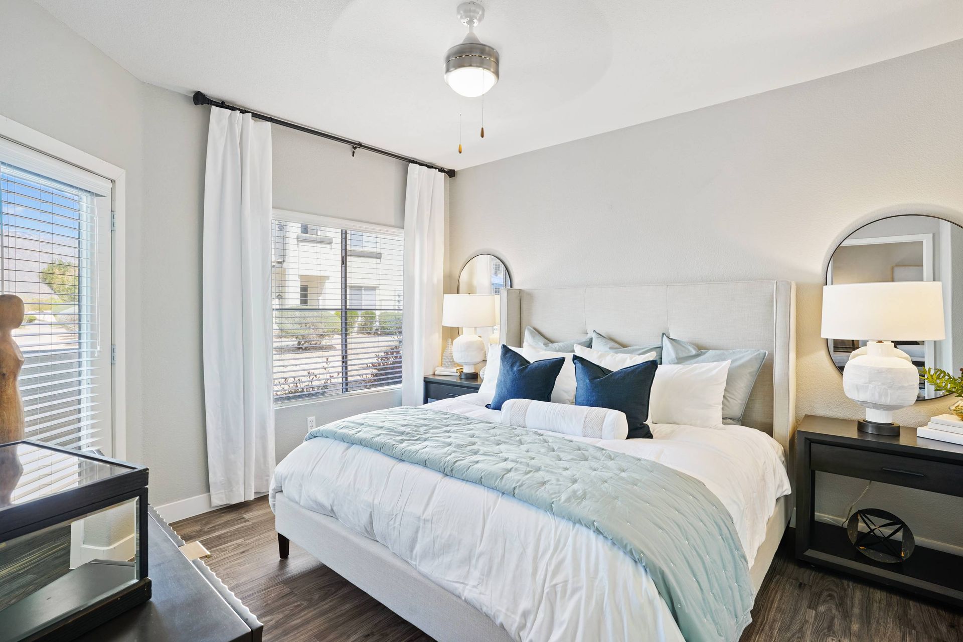 Bedroom in apartment with an upholstered headboard, two nightstands, round mirrors, and a window with white curtains.