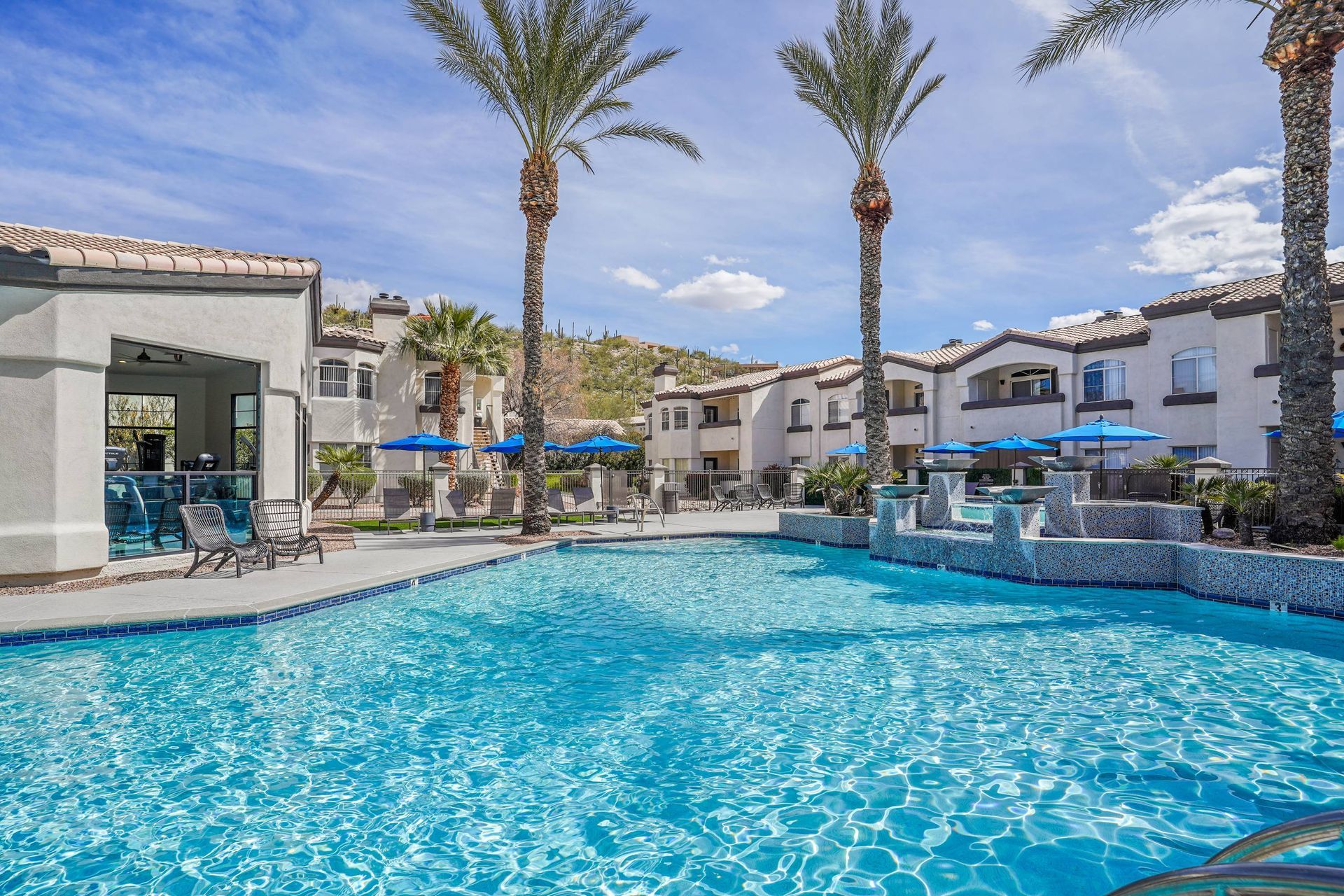 Outdoor community pool with palm trees, blue umbrellas, and lounge seating at a multifamily complex.