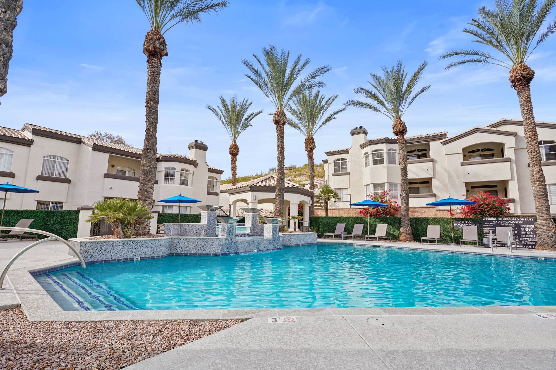 Outdoor pool area with palm trees, lounge chairs, blue water, and surrounding apartment buildings.