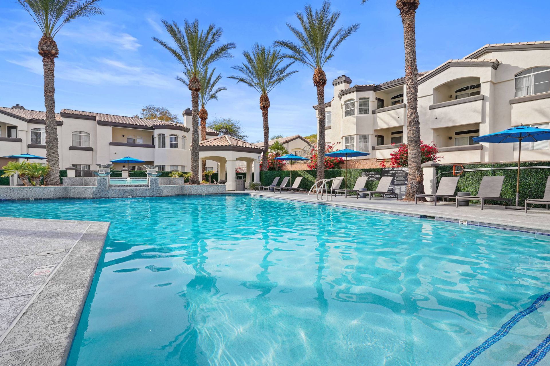 Outdoor pool area with palm trees, lounge chairs, and blue umbrellas at an apartment complex.