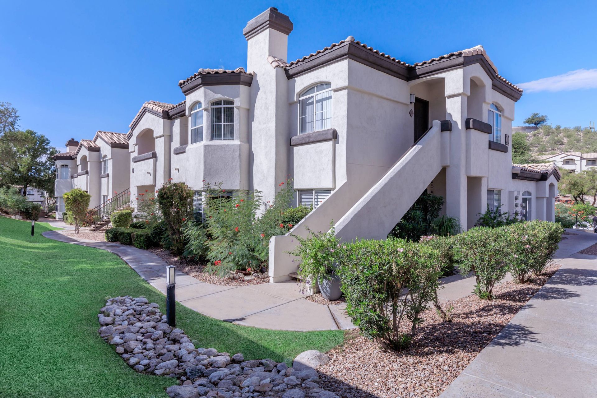 Exterior view of a beige stucco apartment building with a long staircase, windows, and landscaped walkways.