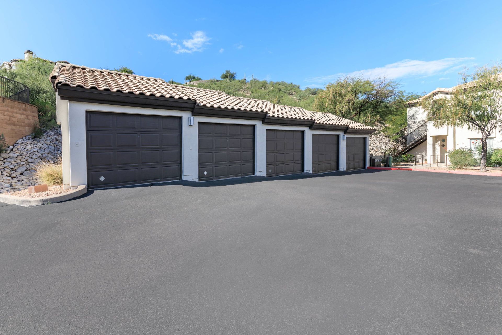 Row of five dark garage doors in a stucco building with a tile roof, a paved lot in front.