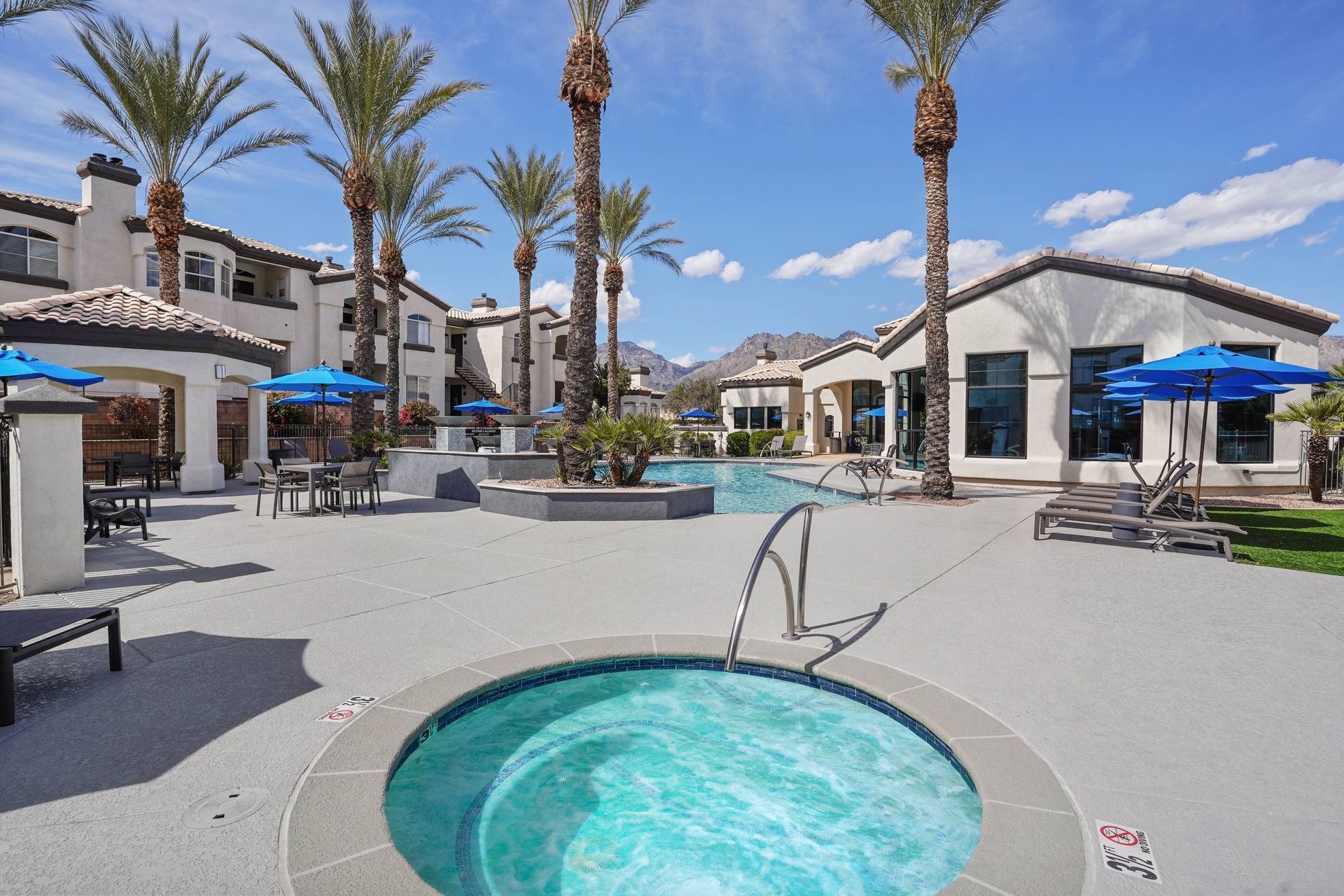 Outdoor pool area with palm trees, lounge chairs, and blue umbrellas at an apartment community.