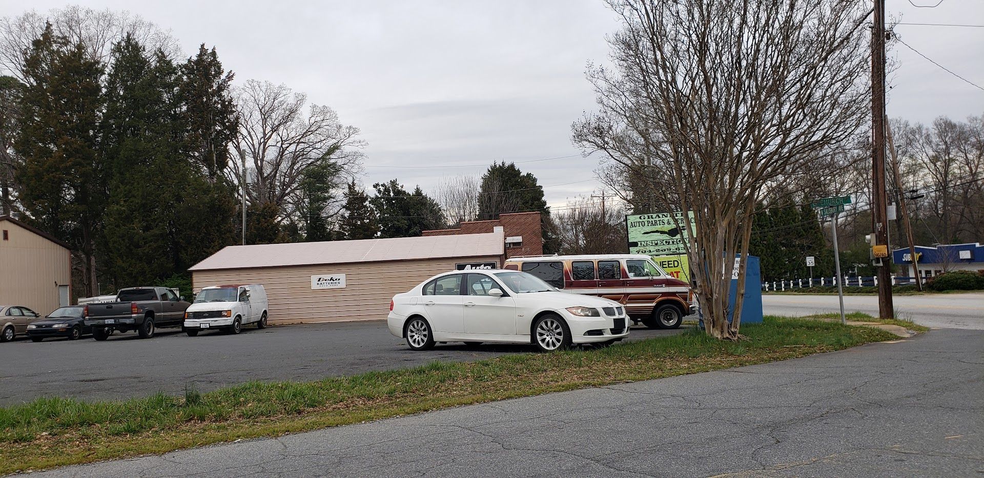 Cars parked in a gravel lot next to a building and tree. Overcast sky | Lowry Auto Service