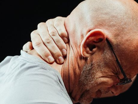 A bald man with glasses is holding his neck in pain.