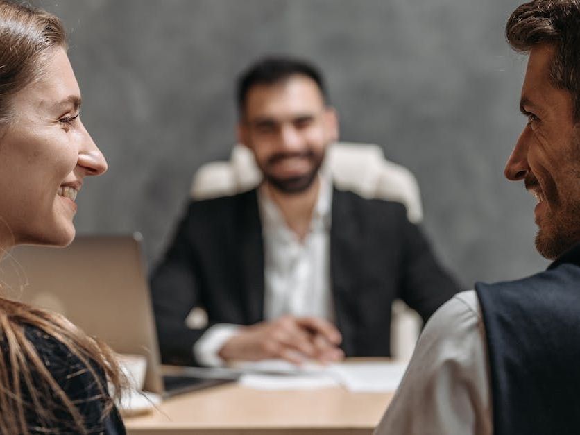 A man and a woman are sitting at a table talking to a man in a suit.