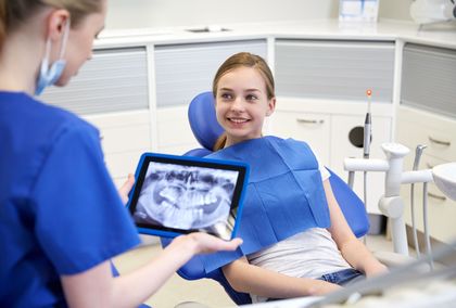 Un dentista in camice blu mostra una radiografia dentale digitale su un tablet a un paziente seduto sulla poltrona odontoiatrica.
