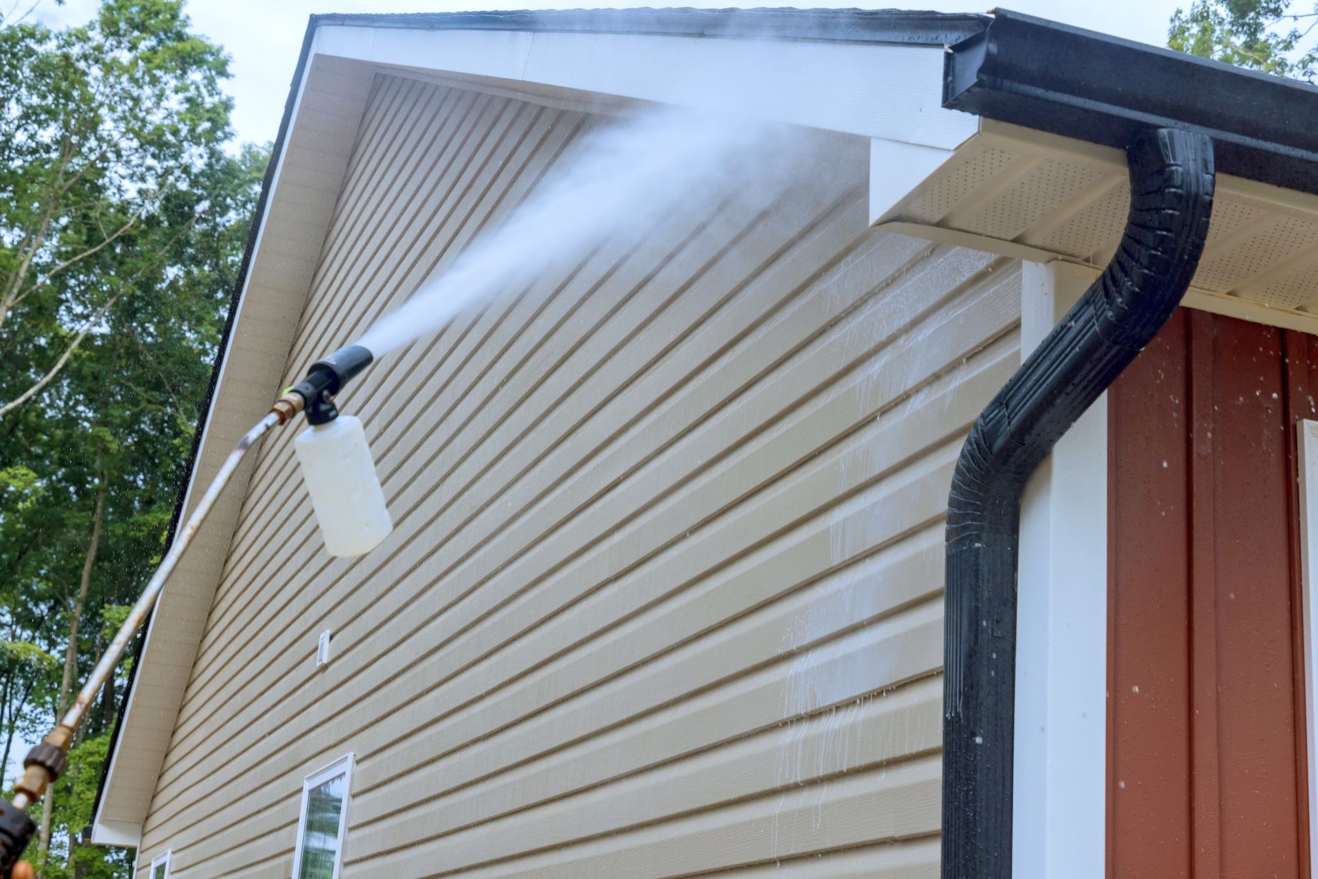 Person pressure-washing beige house siding near the roofline with a spray wand and black gutter.