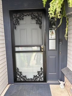 Black ornate screen door with floral detail on a gray front door with a small window and ferns.