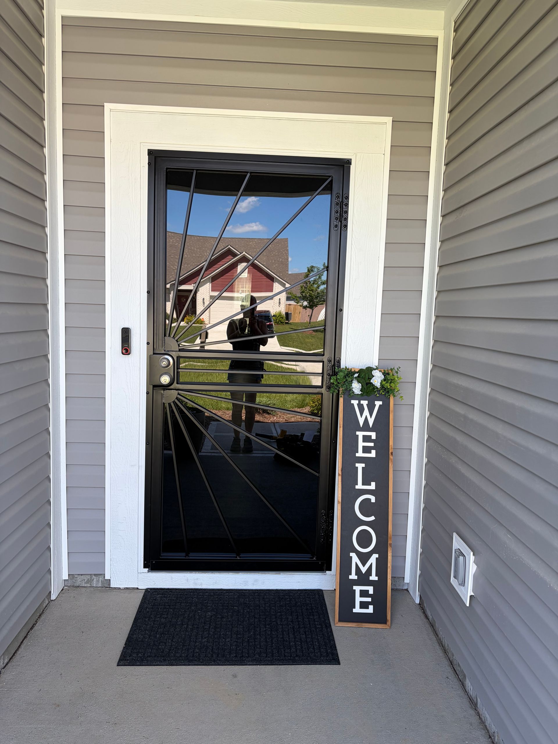 Front porch entry with black door, doormat, and a vertical “WELCOME” sign beside the door.