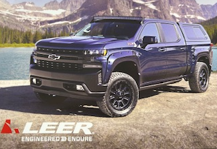 Blue Chevy truck with black rims and camper shell parked by a lake and mountains. Leer logo in the corner.