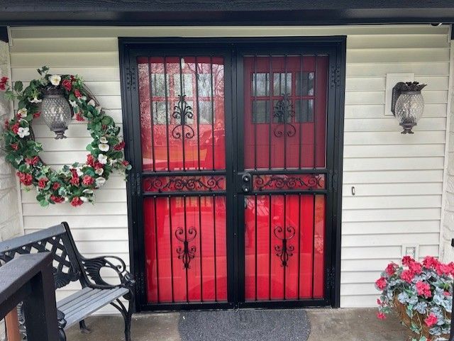 Black security doors with red interior. White siding, floral wreath, and bench.