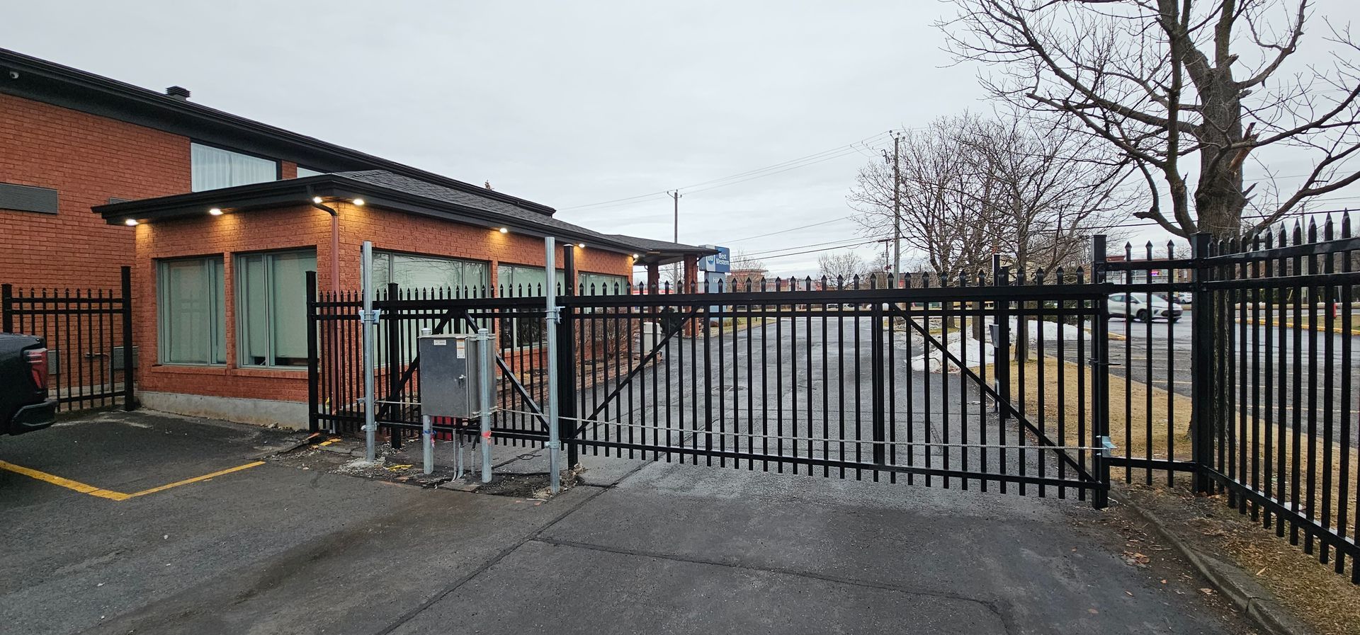 Une clôture noire entoure un parking devant un bâtiment en briques.