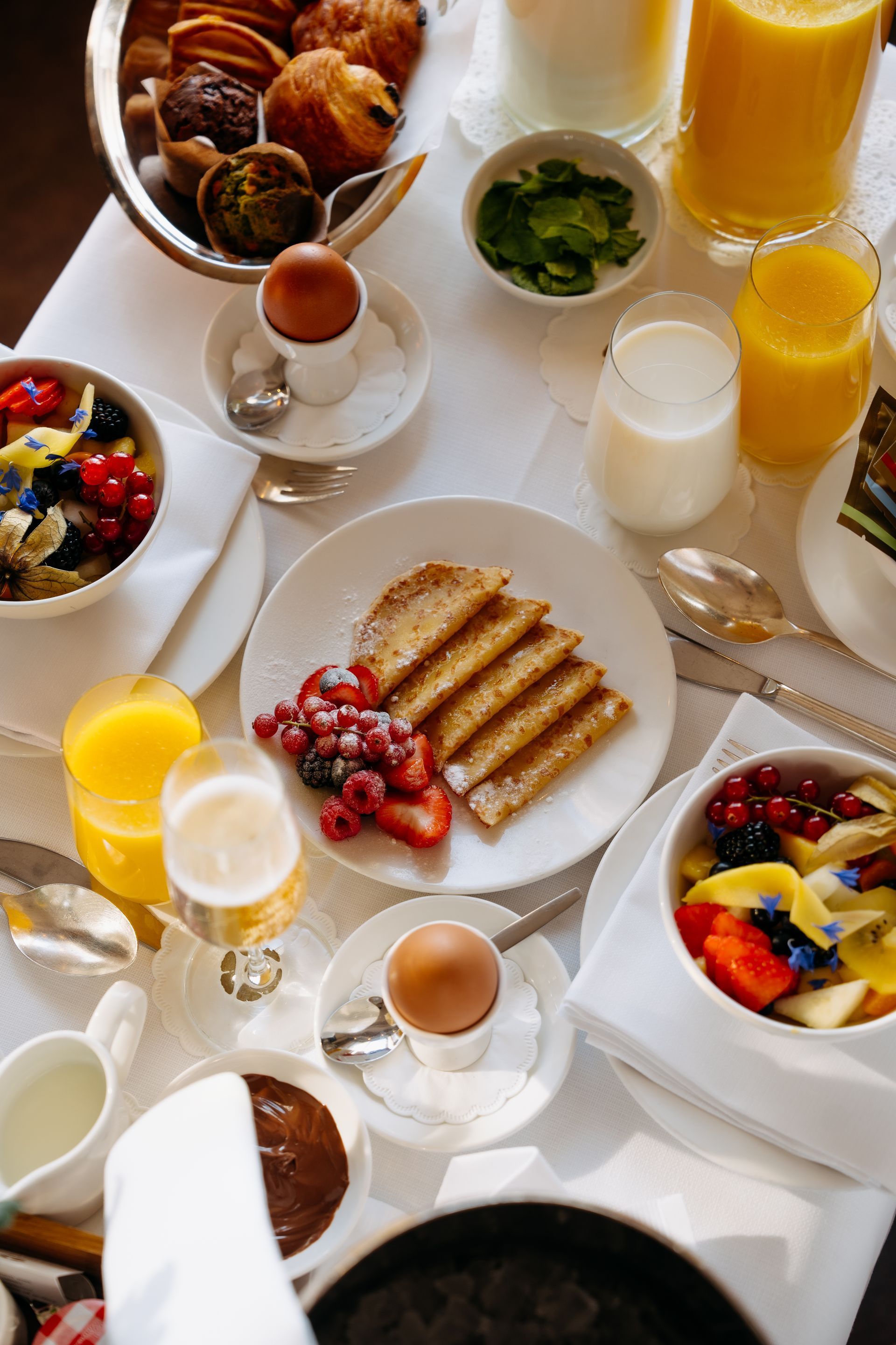 Breakfast spread: toast, fruit, pastries, eggs, orange juice, and milk on a white tablecloth.