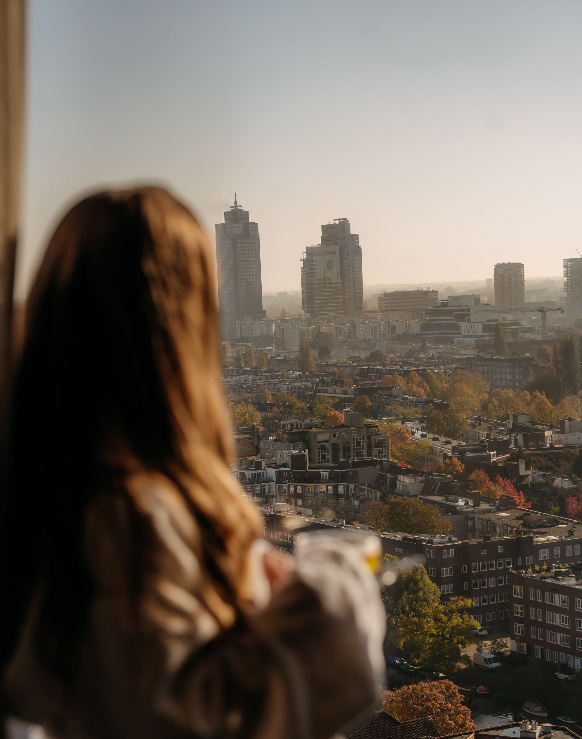 Woman looking out a window at a hazy city view, holding a drink.