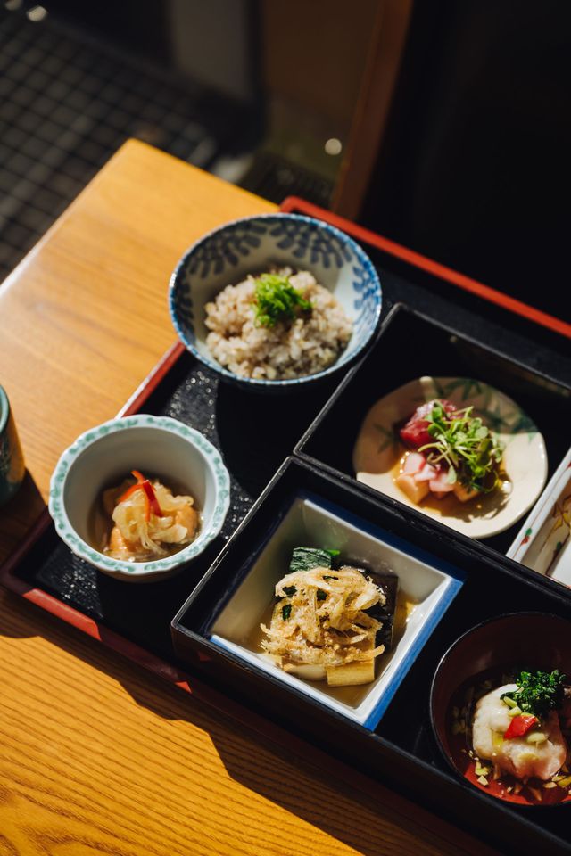 Bento box with various Japanese dishes, including sushi, seafood, and salad, presented on a dark surface.
