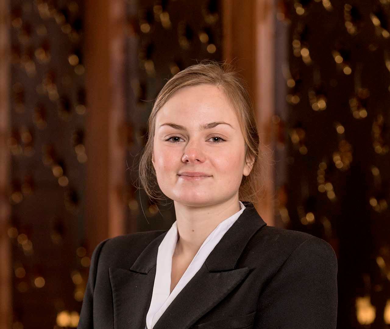Woman in black blazer with white shirt, smiling, in front of a patterned brown backdrop.