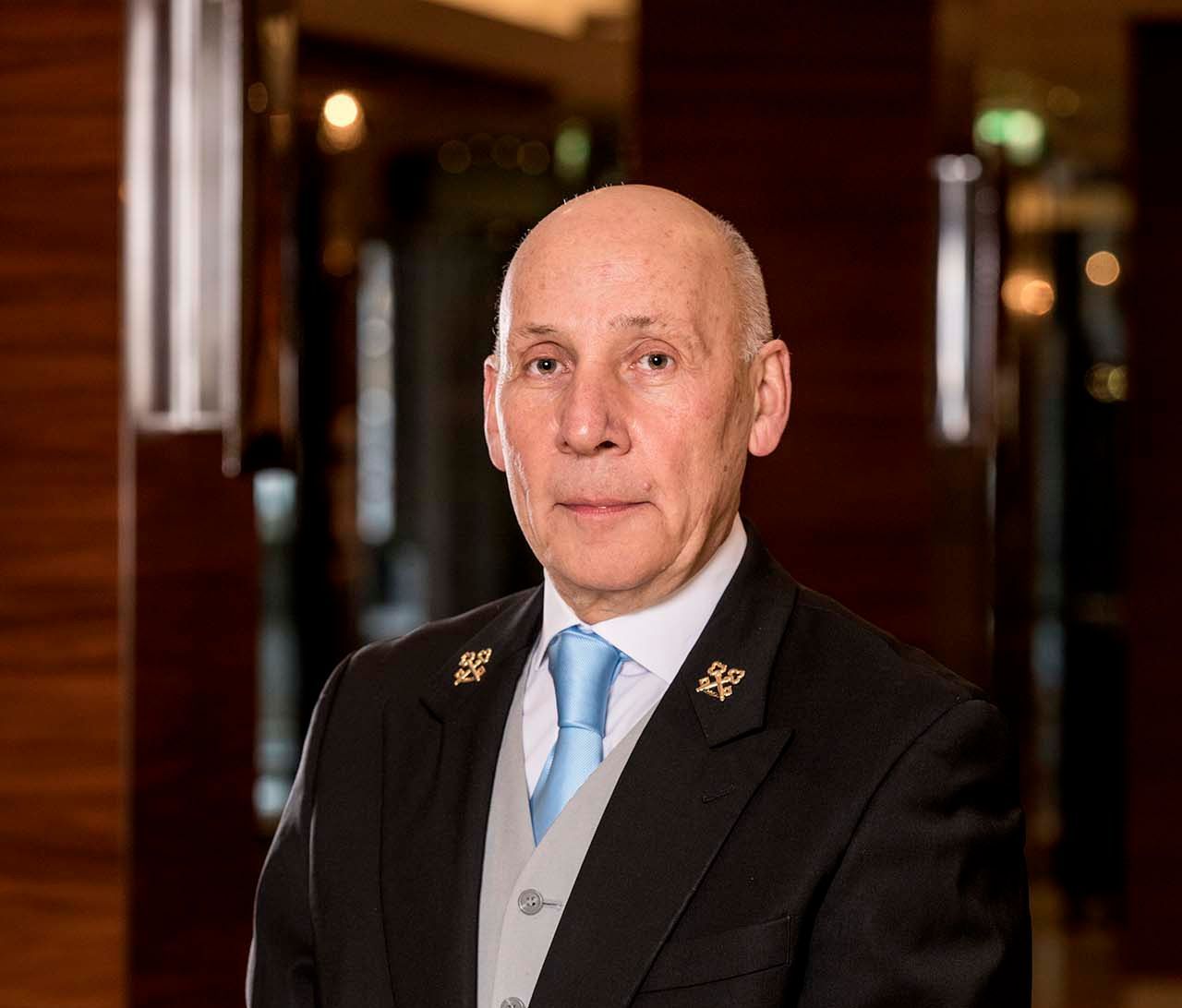 Man in formal attire, light blue tie, black suit, and gold emblems, posing indoors.