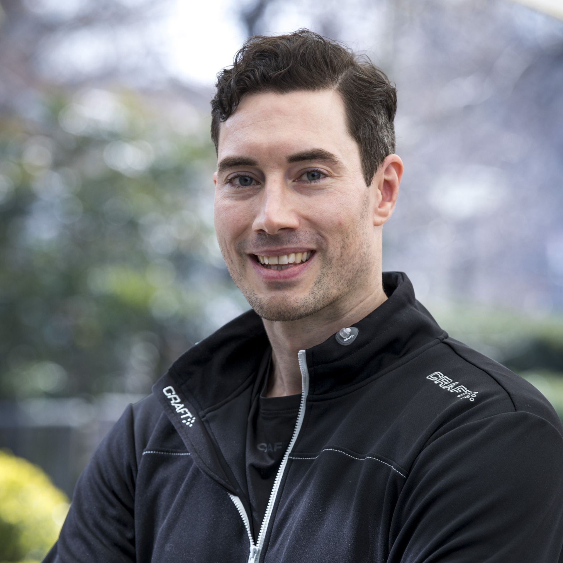 Man in black zip-up jacket smiles in an outdoor setting.
