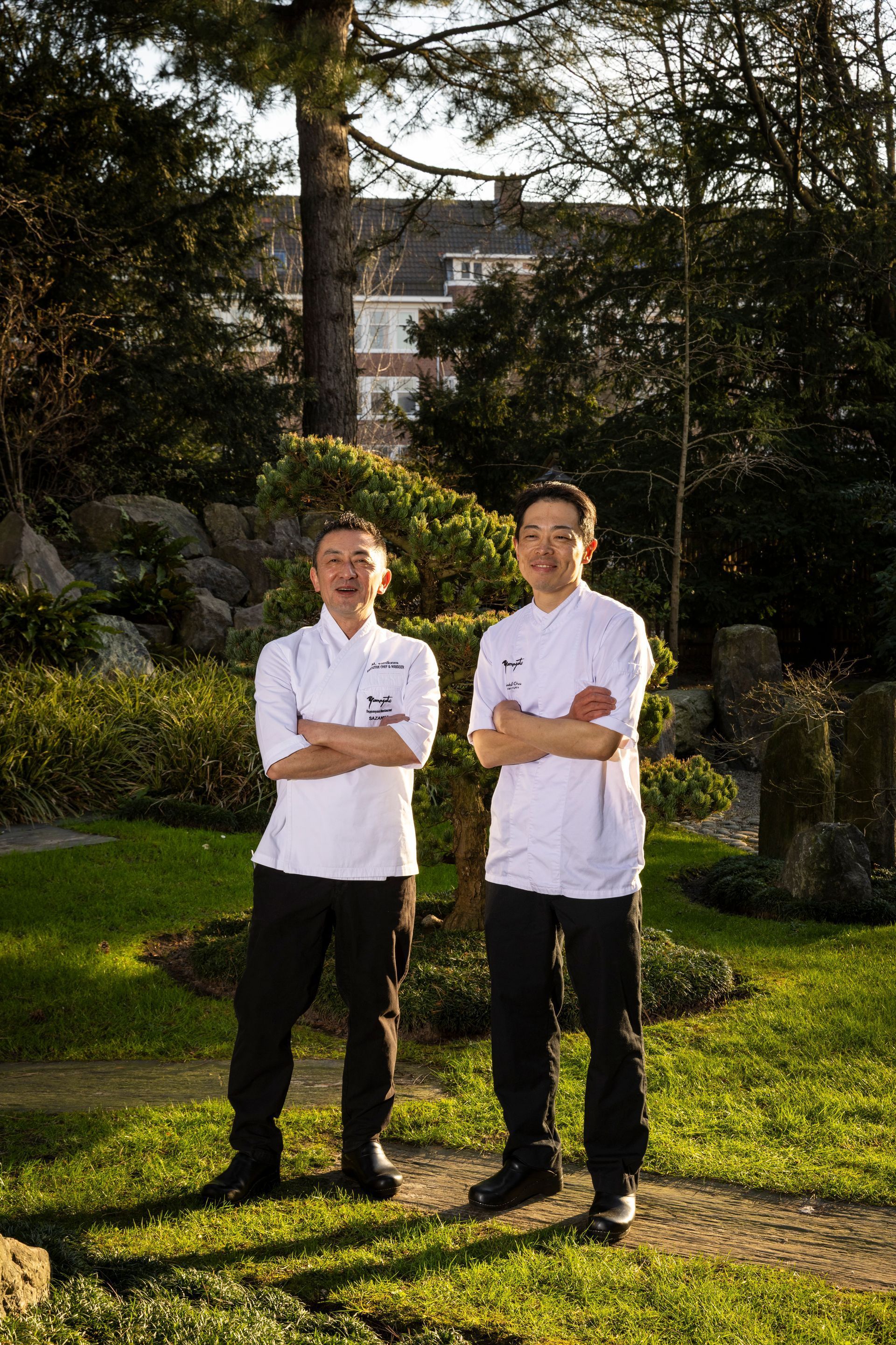 Two chefs in white coats with crossed arms standing in a garden.
