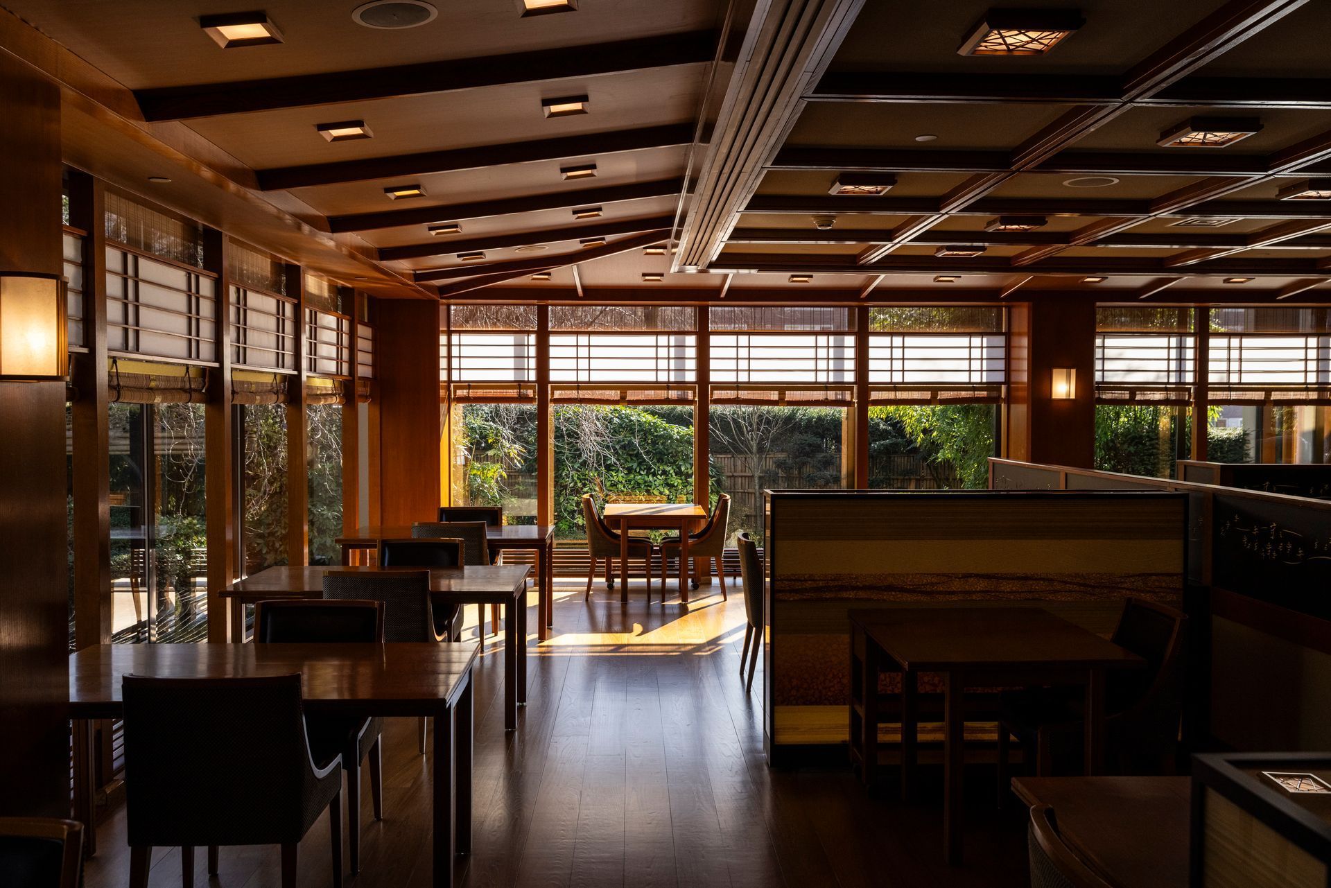 Empty, wood-paneled dining room with large windows, tables, and chairs. Sunlight streams in.