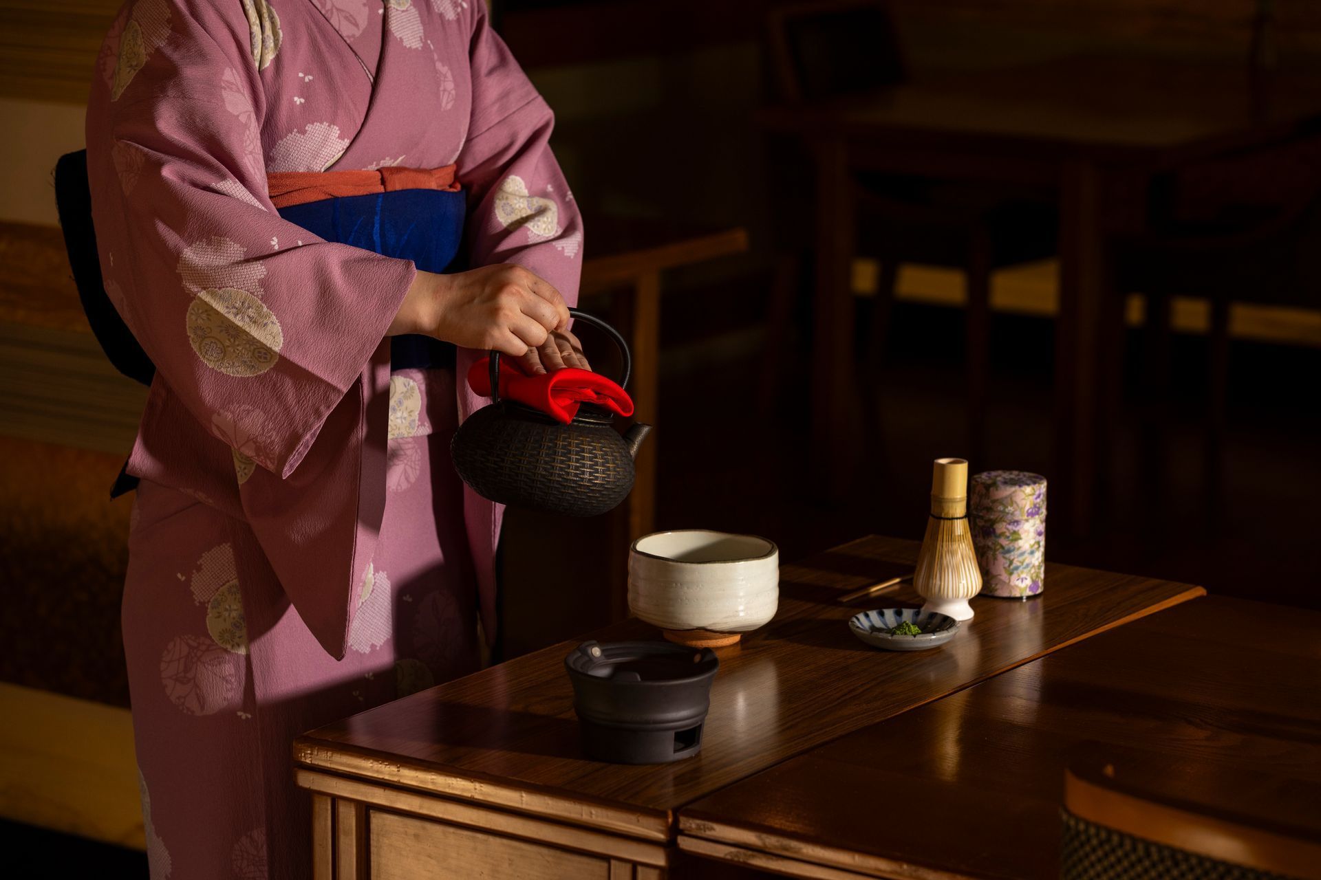 Woman in kimono pouring tea into a ceramic bowl, preparing a traditional tea ceremony.