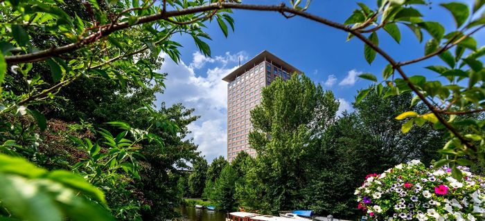 A boat on a river in front of a tall building framed by lush green foliage and flowers.