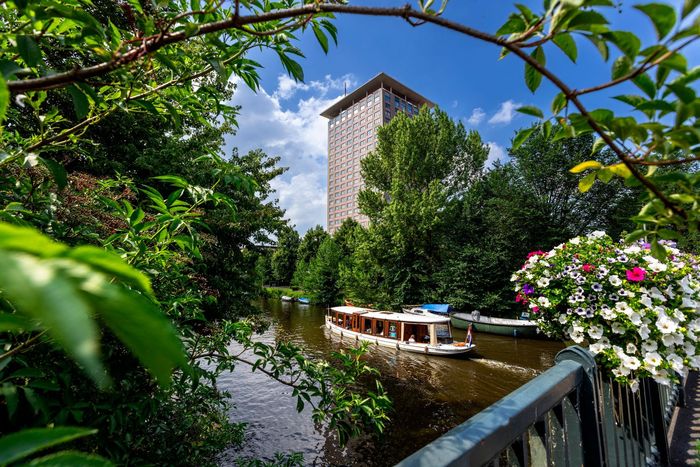 A boat on a river in front of a tall building framed by lush green foliage and flowers.