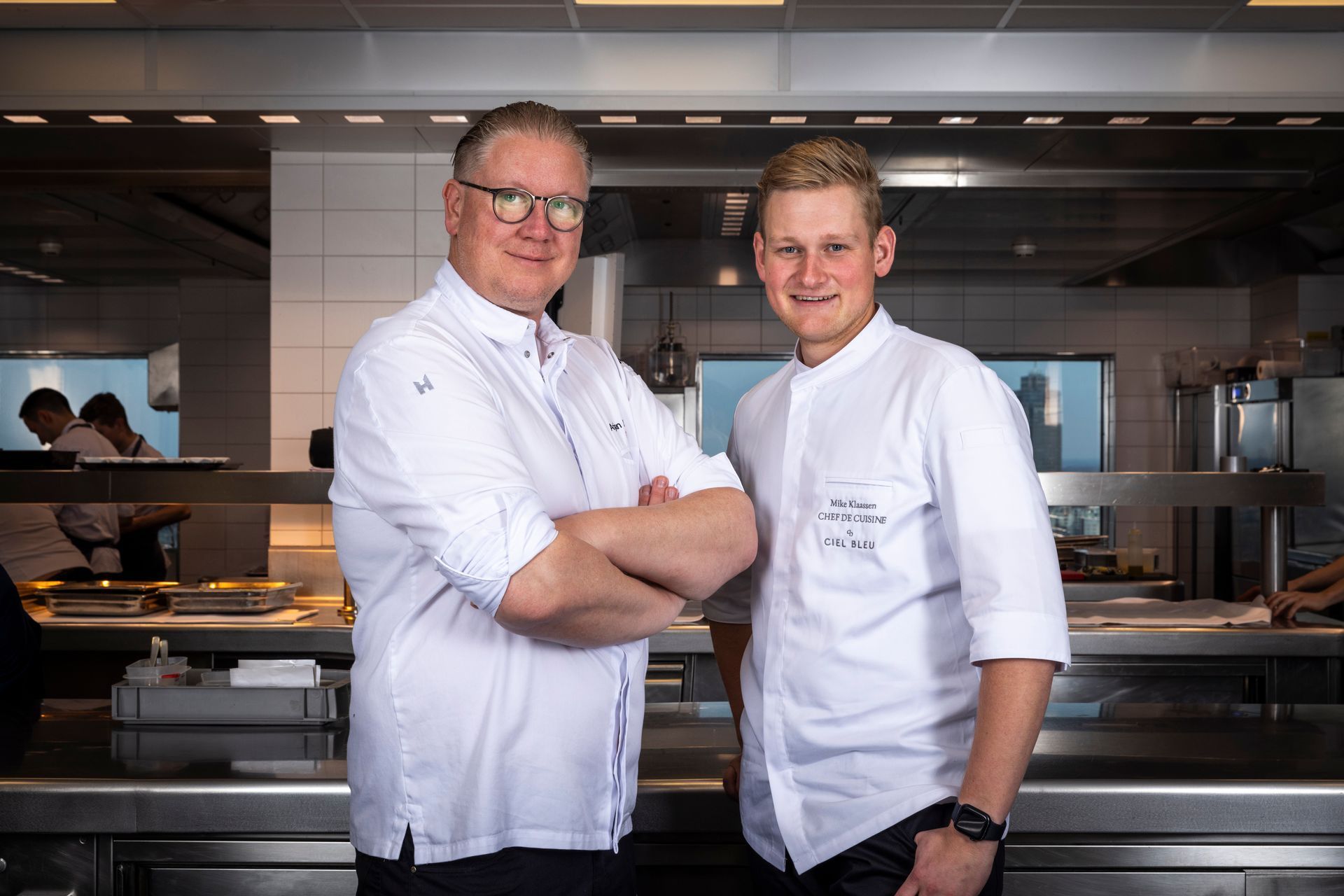 Two chefs in a kitchen; one with crossed arms, the other smiling. Stainless steel counters.
