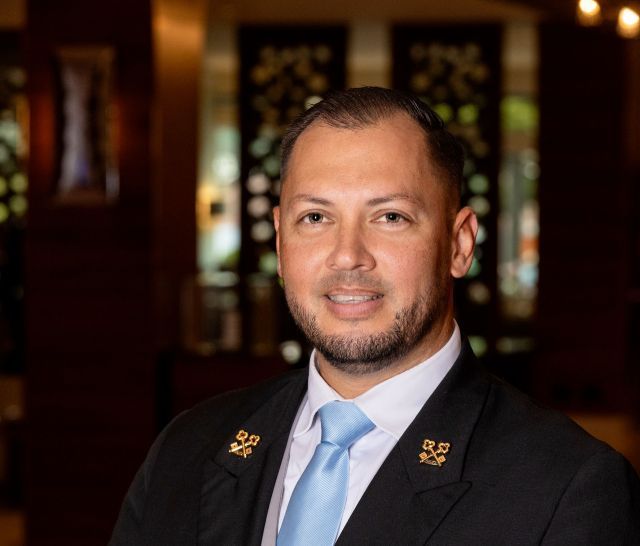 Man in suit and tie smiling, standing indoors, hotel setting.