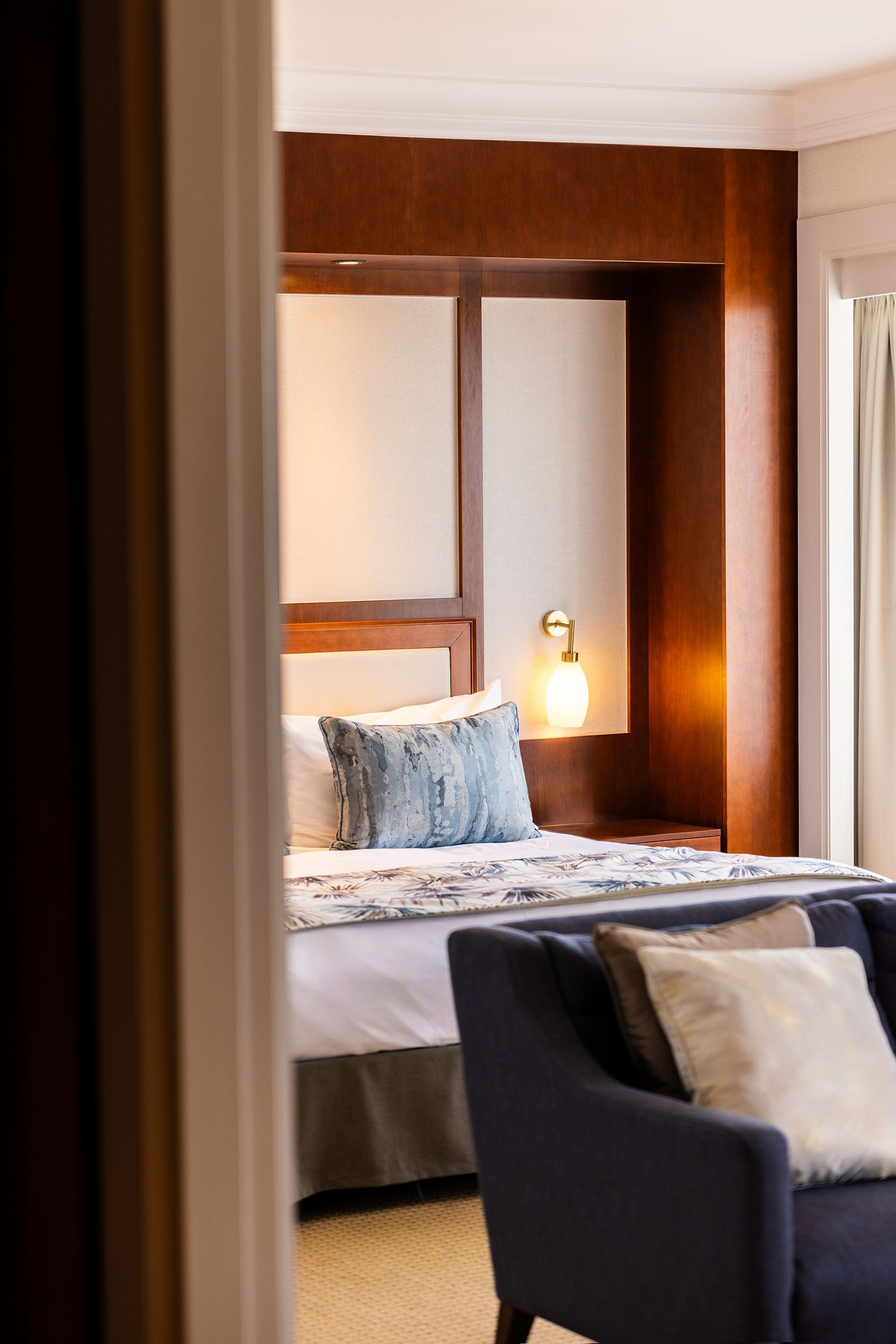 Bedroom interior, viewed through a doorway. Bed with decorative pillows, lamp, and dark blue couch visible.