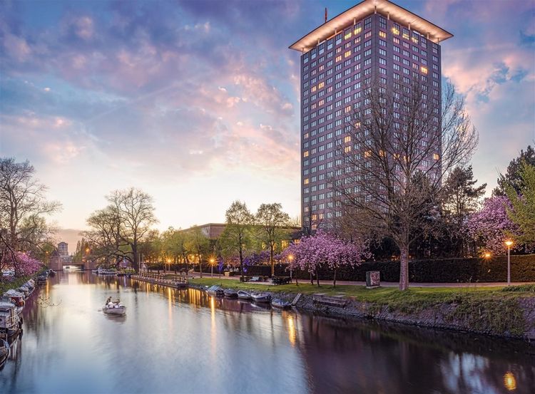 A tall building overlooking a river with a boat. Trees with pink blossoms and twilight sky.