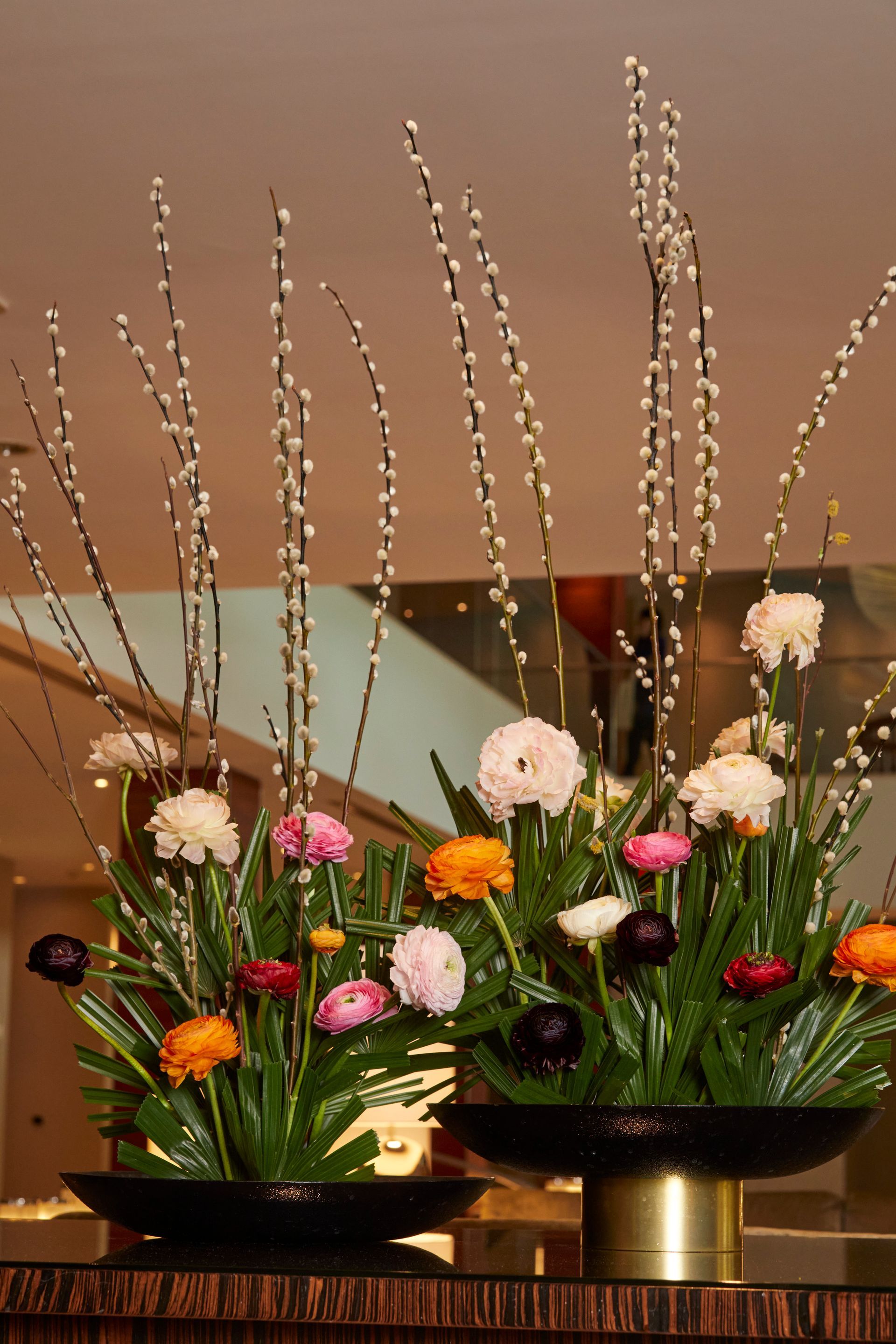 Two floral arrangements with tall, willow branches and colorful blooms in black bowls.