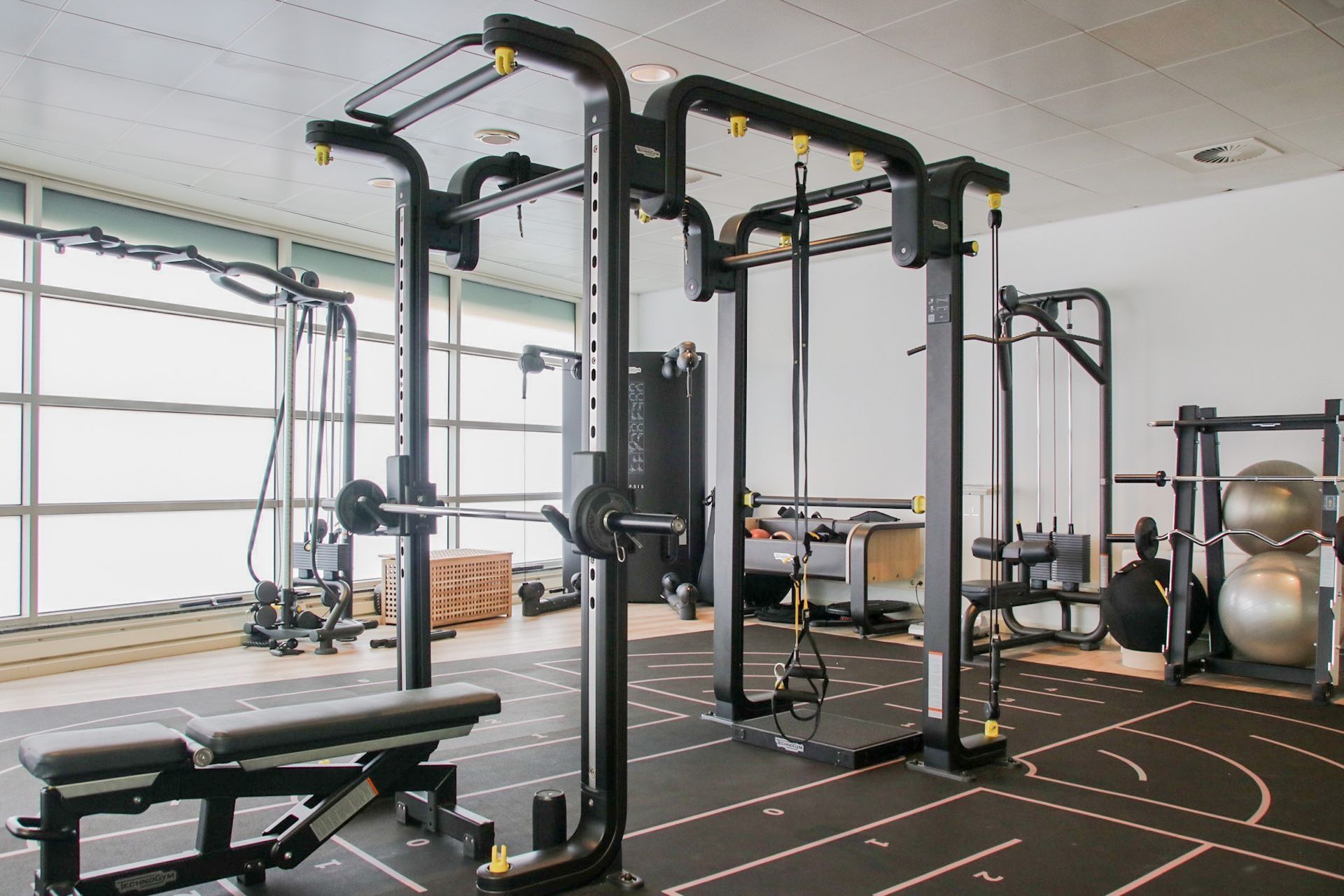 Gym interior with weightlifting equipment; power rack, cable machine, bench, barbells, and floor markings.